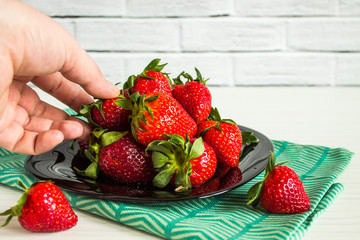 Bright ripe red strawberries in a black plate with green textiles close up on a white brick background with copy space and a human hand