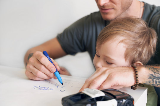 Young father helps son at age of two draw, teaches alphabet. A small boy is sitting on his dads lap near white wall in room at table, on piece of paper writing letters and pictures with felt-tip pen.