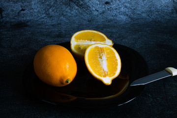 A whole lemon and two halves on a black plate and a kitchen knife on a grey textured background in...