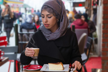 Portrait of young Muslim woman drinking coffee at her hand in cafe. She is wearing a turban (hijab, head scarf) and she is sitting outside.