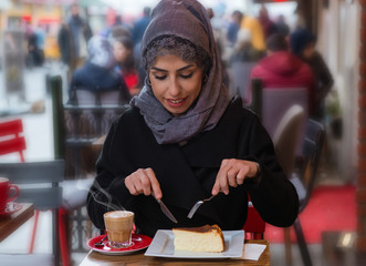 Portrait of young Muslim woman eating san sebastian cheseecake with cup of hot coffee. She is wearing a turban (hijab, head scarf) and she is sitting outside.