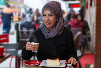 Portrait of young Muslim woman drinking coffee at her hand in cafe. She is wearing a turban (hijab, head scarf) and she is sitting outside.