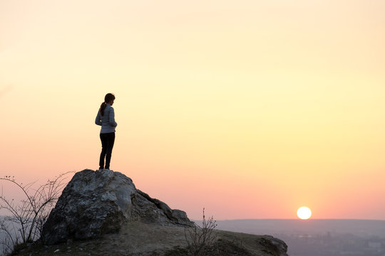 Silhouette of a woman hiker standing alone on big stone at sunset in mountains. Female tourist on high rock in evening nature. Tourism, traveling and healthy lifestyle concept.