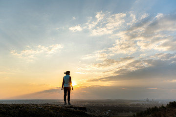 Silhouette of a woman hiker standing alone enjoying sunset outdoors. Female tourist on rural field in evening nature. Tourism, traveling and healthy lifestyle concept.