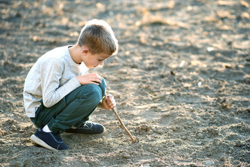Child boy playing with wooden stick digging in black dirt ground outdoors.