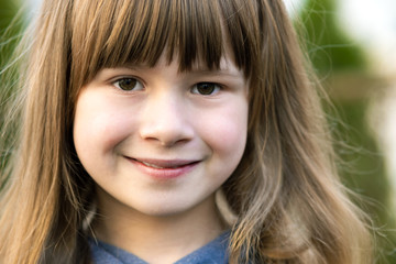 Portrait of pretty child girl with gray eyes and long fair hair smiling outdoors on blurred green bright background. Cute female kid on warm summer day outside.