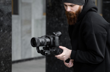 Bearded Professional videographer in black hoodie holding professional camera on 3-axis gimbal...