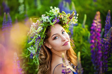 a young beautiful girl stands on a blooming spring meadow of lupines with a flower wreath on her head at the sunset of spring.
