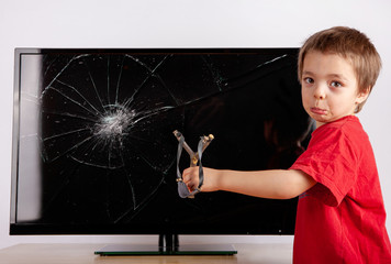 Shocked little boy standing in front of a TV with broken screen and showing his slingshot. Home...