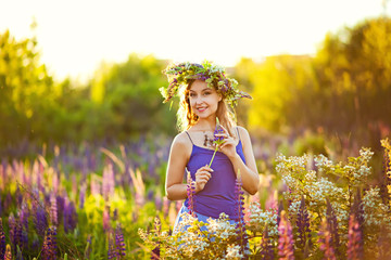 a young beautiful girl stands on a blooming spring meadow of lupines with a flower wreath on her head at the sunset of spring.