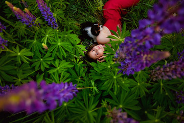 A girl in a clearing with lupine flowers in the forest, holding a small dog Cavalier king Charles Spaniel, a wreath of flowers on her head.