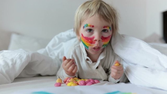 Beautiful Blond Toddler Boy With Rainbow Painted On His Face And Messy Hands, Smiling Happily