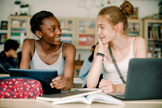 Smiling teenagers talking while sitting by table in classroom