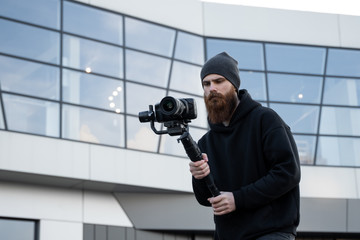 Bearded Professional videographer in black hoodie holding professional camera on 3-axis gimbal...