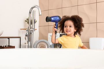 Little African-American girl playing with blow dryer in bathroom. Child in danger