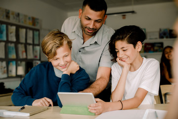 Smiling male tutor with students using digital tablet while sitting in classroom
