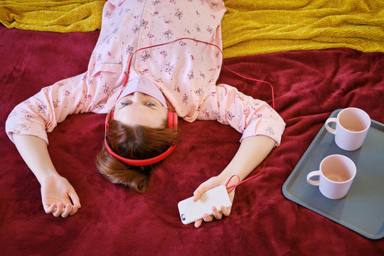 A Woman In A Makeshift Medical Mask Is Isolated During Quarantine By The Coronavirus. The Girl On The Bed Listens To Music From The Phone In The Headphones