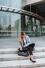 A young attractive businesswoman with a bag is sitting on the stairs in front of the company