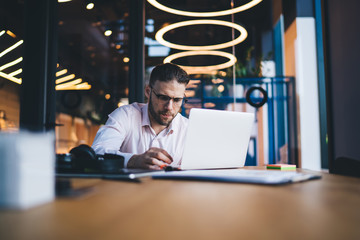 Intelligent male software developer in optical glasses typing program code during remote working on freelance, skilled businessman booking tutorial webinar on sale connecting to 4g wireless on laptop