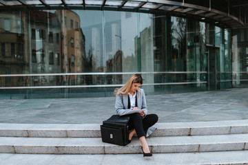 A young attractive businesswoman with a bag is sitting on the stairs in front of the company
