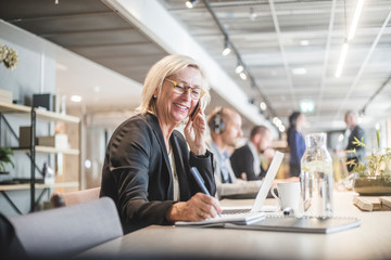 Smiling businesswoman talking on mobile phone and writing in diary while working at office desk