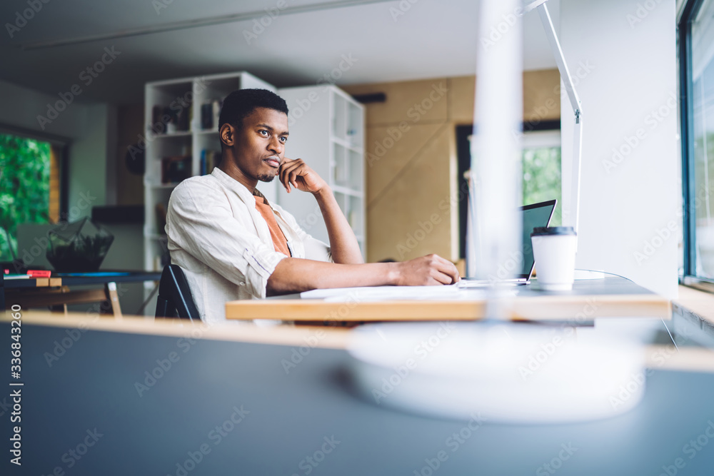 Poster millennial programmer sitting at table desktop in coworking space and pondering on information while