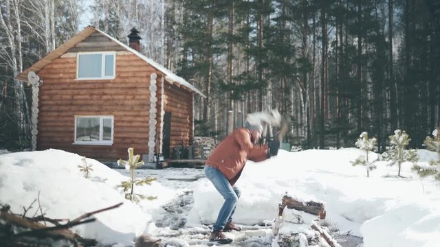 A Caucasian man in a brown jacket, hat, jeans and boots, holding an axe, chopping wood in winter. A man stands in the courtyard of a country house, which is located next to the forest.