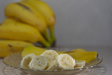 Banana (Musa ssp.) Served in slices on a glass plate and in the background a bunch of defocused bananas