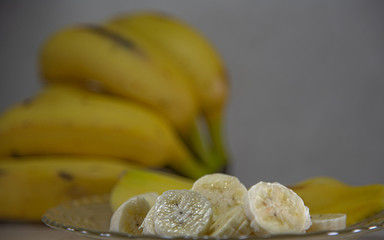 Banana (Musa ssp.) Served in slices on a glass plate and in the background a bunch of defocused bananas