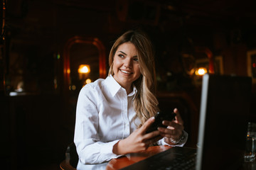 A young attractive caucasian  blond girl with a phone is sitting in a cafe in front of a laptop