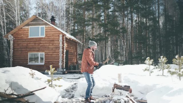 A Caucasian man in a brown jacket, hat, jeans and boots, holding an axe, chopping wood in winter. A man stands in the courtyard of a country house, which is located next to the forest.