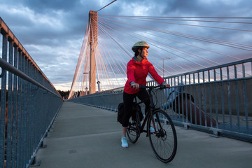 Caucasian Woman riding a Bicycle on a path at Port Mann Bridge during vibrant Sunset. Taken in Surrey, Vancouver, British Columbia, Canada.