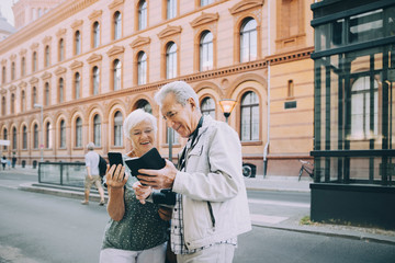 Smiling senior male and female tourist using smart phone while exploring in city