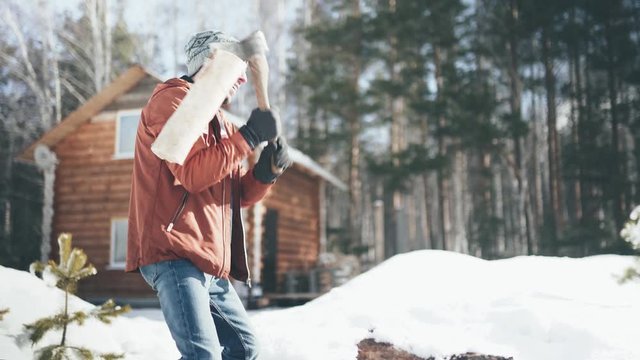 A Caucasian man in a brown jacket, hat, jeans and boots, holding an axe, chopping wood in winter. A man stands in the courtyard of a country house, which is located next to the forest.