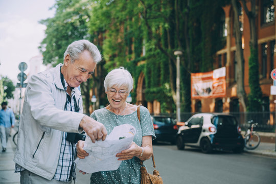 Smiling elderly couple reading map while standing on sidewalk in city during vacation