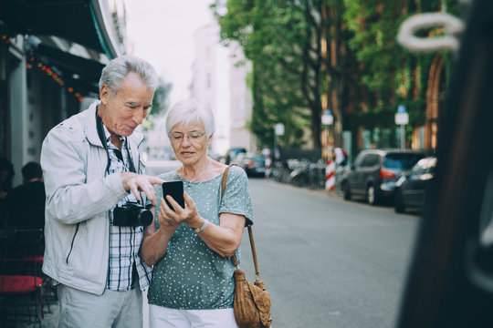 Senior male and female using smart phone while standing on sidewalk in city