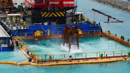 Fototapeta na wymiar Huge excavator on floating platform in the blue water of the Pacific Ocean extract white sand from the bottom and load onto a barge amid clouds and tropical islands. close up crane basket plunge