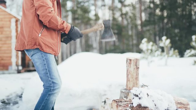 A Caucasian man in a brown jacket, hat, jeans and boots, holding an axe, chopping wood in winter. A man stands in the courtyard of a country house, which is located next to the forest.