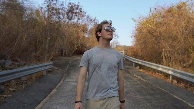 Follow Shot of Handsome Young Man Walking Downhill on Empty Road in Mountains During Summer