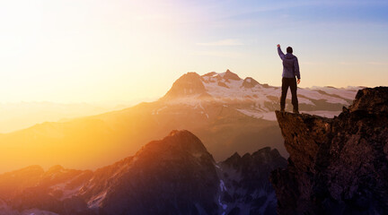 Composite. Adventurous Man Hiker With Hands Up on top of a Steep Rocky Cliff. Sunset or Sunrise. Landscape Taken from British Columbia, Canada. Concept: Adventure, Explore, Hike, Lifestyle