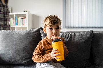 Portrait of small caucasian boy three or four years old sitting on the bed or sofa at home holding a plastic cup with straw drinking juice or soda at home alone in day looking to the camera