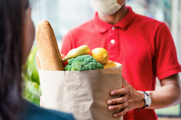 Asian deliver man wearing face mask in red uniform handling bag of food, fruit, vegetable give to...