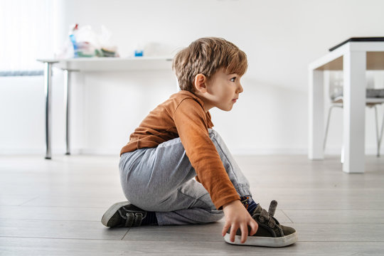 Full length view of small cute caucasian boy little child kid sitting on the wooden or laminated vinyl floor at home putting on or taking off the shoes in day alone side view