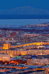 Aerial landscape with the northern city. Beautiful evening cityscape with many buildings. Bright street lighting at dusk. In the distance are mountains and a sea bay. Magadan, Siberia, Far East Russia