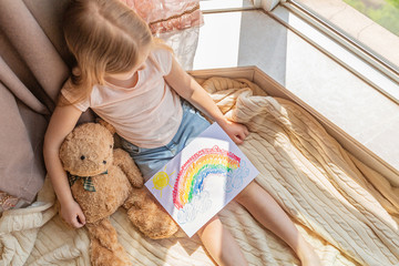 Kid with painted rainbow during Covid-19 quarantine at home. Caucasian Girl with teddy bear toy...