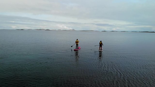 4K aerial drone halv orbit right motion of two people stand up paddling into the sea. Amazing Northern Norway landscape