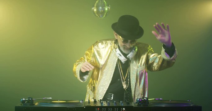 Funny retro style Caucasian old man in golden jacket djing at nightclub. Vintage senior dj dancing while mixing music on audio equipment. Stylish clubbing grandpa having fun at party. Disco concept