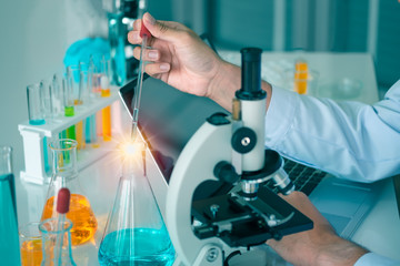 Close-up shot of the hand of a male scientist is experimenting with tube and microscope. Laboratory there is glassware equipment that different from liquids and laptop on the table for research.