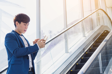 Asian handsome young business smiling and holding smartphone while standing on the escalator. Business and finance concept.