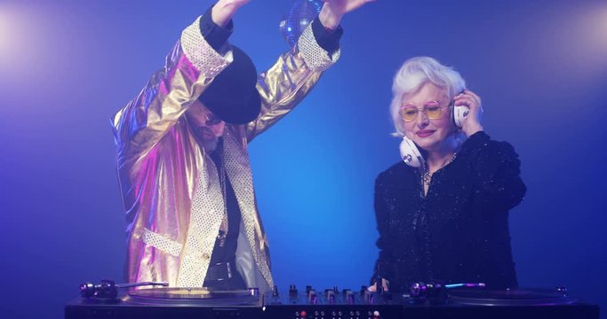 Funny vintage stylish couple of old male and female DJs playing disco at party. Dancing retro style Caucasian senior man in sunglasses and elder lady with headphones near vinyl system Clubbing concept